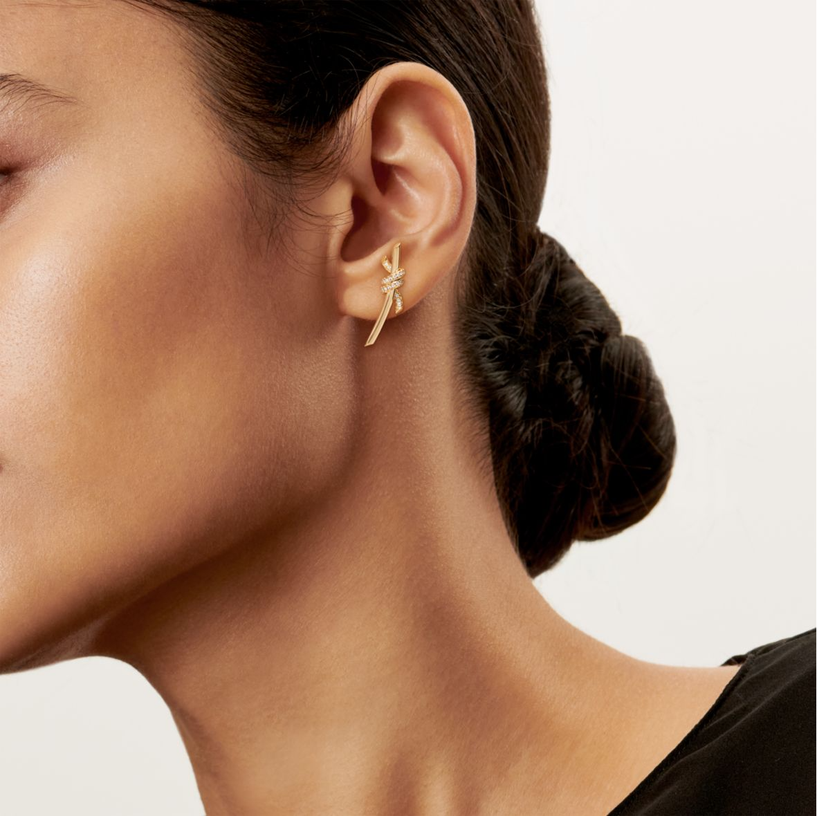 Close-up of an ear wearing a gold earring with a plain background Inspired by the timeless elegance of classic design, the Knot of Love Earrings embody the strength of unbreakable bonds and cherished connections. Each piece is meticulously crafted in 18k yellow gold and adorned with hand-set round brilliant diamonds, chosen for their exceptional sparkle and precision. The design symbolizes unity, making these earrings a modern expression of love, devotion, and enduring beauty.