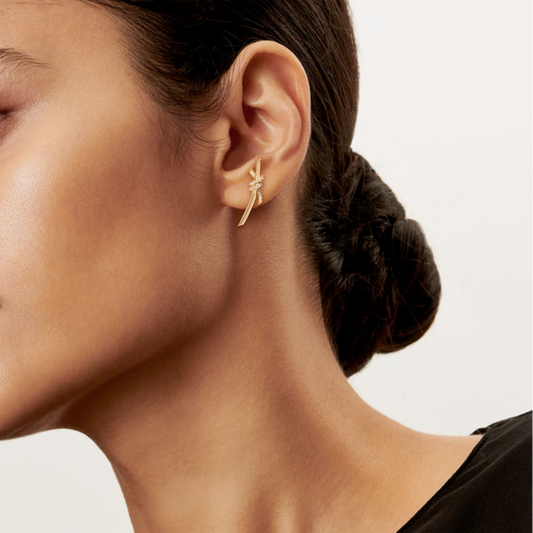 Close-up of an ear wearing a gold earring with a plain background Inspired by the timeless elegance of classic design, the Knot of Love Earrings embody the strength of unbreakable bonds and cherished connections. Each piece is meticulously crafted in 18k yellow gold and adorned with hand-set round brilliant diamonds, chosen for their exceptional sparkle and precision. The design symbolizes unity, making these earrings a modern expression of love, devotion, and enduring beauty.